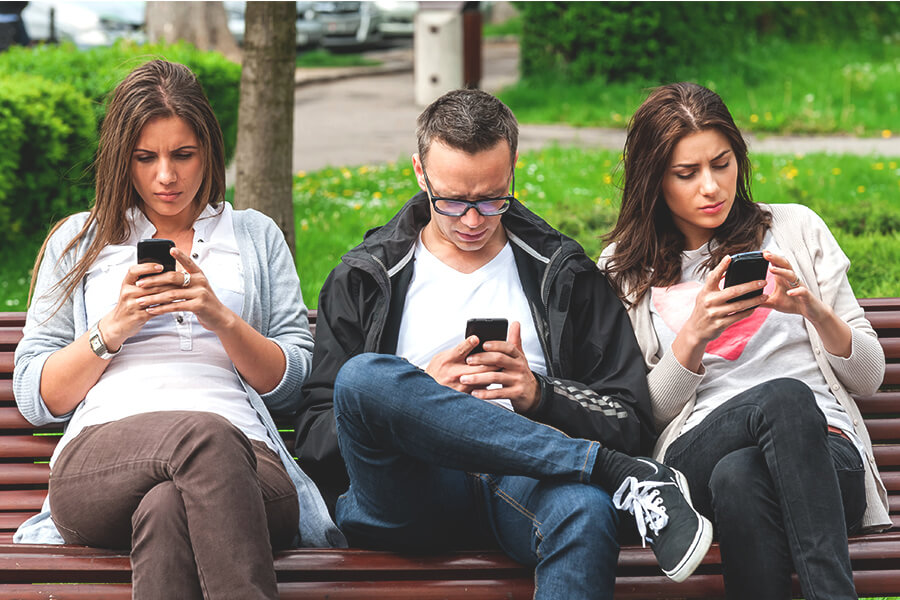 Three people sitting on a park bench looking at their phones with forward head posture