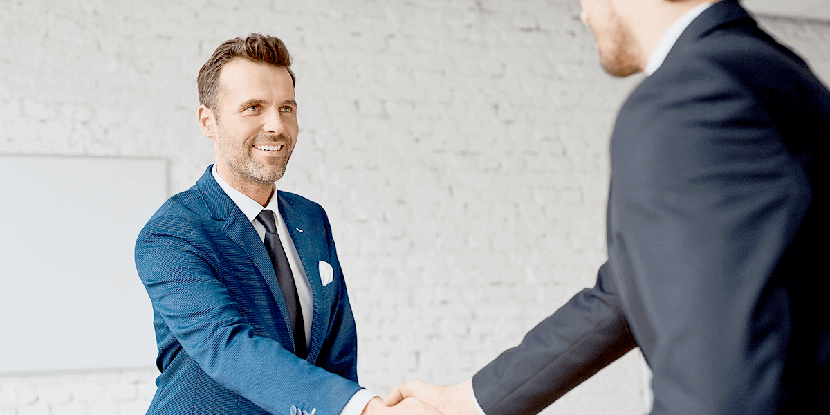 Two businessmen smiling and shaking hands in front of a white wall